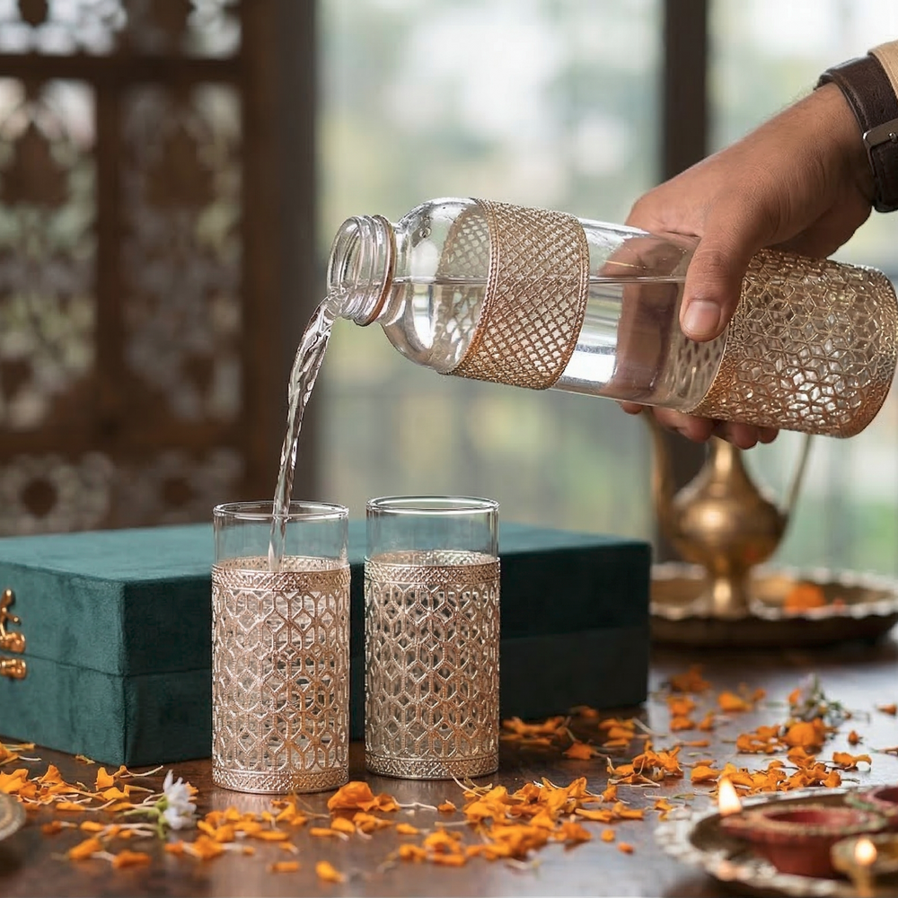 Person pouring liquid from a decorative bottle into two glasses on a table with flowers and a box.