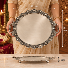 A person in a beige embroidered dress holds an oval silver-toned decorative tray with ornate filigree edges and side handles. The image includes dimension annotations showing the tray is 38 cm by 38 cm, with a smaller view of the tray on a marble surface below.