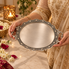 A person in a beige and gold embroidered traditional outfit holds an oval silver tray with ornate filigree borders and decorative handles. The scene is set on a marble tabletop with flowers, a lit candle holder, and warm ambient lighting.