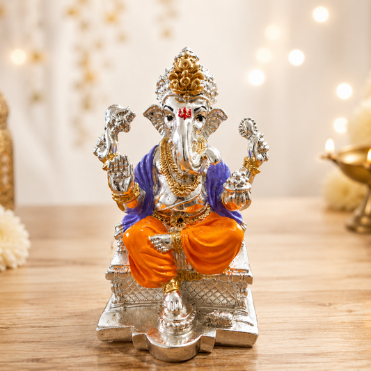 A silver-toned Lord Ganesha idol with orange and purple draped garments, seated on a decorative base on a wooden table, surrounded by festive decor including marigold flowers, a brass lantern, and a diya with warm blurred lights in the background.