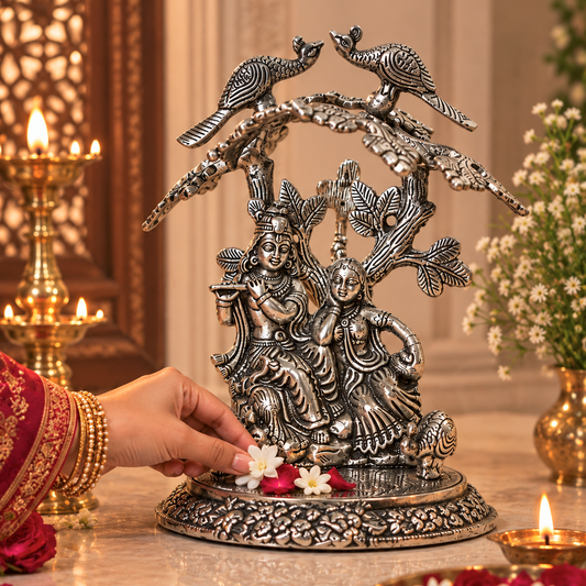 An ornate silver-toned metal sculpture of Lord Krishna and Rukmini under peacock birds, displayed on a marble altar with a hand placing flowers at its base, surrounded by lit oil lamps and decorative floral arrangements.