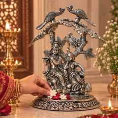 An ornate silver-toned metal sculpture of Lord Krishna and Rukmini under peacock birds, displayed on a marble altar with a hand placing flowers at its base, surrounded by lit oil lamps and decorative floral arrangements.