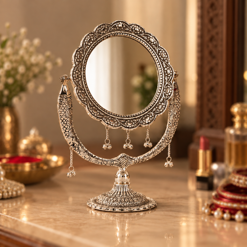An ornate silver-toned vanity mirror with intricate filigree detailing and dangling tassel accents, displayed on a polished marble surface in a warm, decorated interior setting.