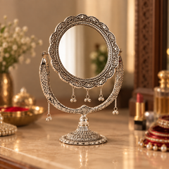An ornate silver-toned vanity mirror with intricate filigree detailing and dangling tassel accents, displayed on a polished marble surface in a warm, decorated interior setting.