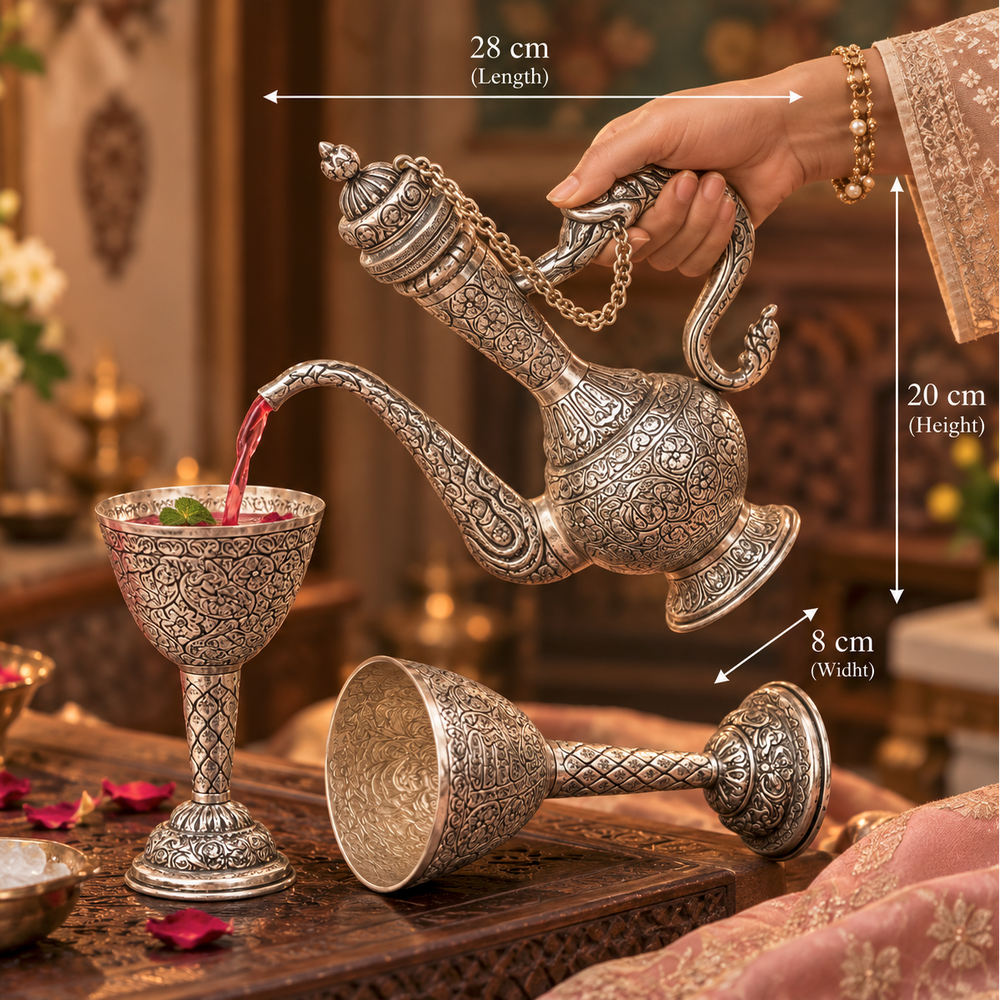 An ornate silver-toned Arabic-style teapot with intricate engraved patterns is being used to pour pink liquid into a matching engraved goblet. A second matching goblet lies on its side below.