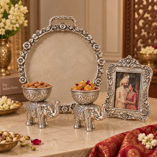 An ornate silver-toned decorative set on a marble tabletop, featuring a large oval tray with a floral border, two elephant-shaped bowls holding sweets, and a matching rectangular picture frame with an embossed portrait. The scene is styled indoors with flowers, a red patterned cloth, and decorative objects in the background.