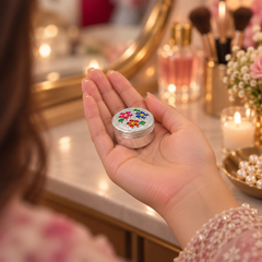 A hand holding a small round silver tin decorated with colorful floral patterns and a rhinestone-embellished lid, set against a softly blurred vanity background with makeup brushes, perfume bottles, and a mirror.