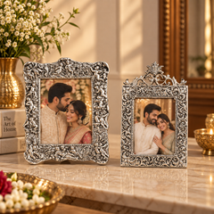Two ornate silver-toned decorative picture frames with intricate scrollwork, displayed on a marble surface in a warm, elegant interior setting with flowers and candles.