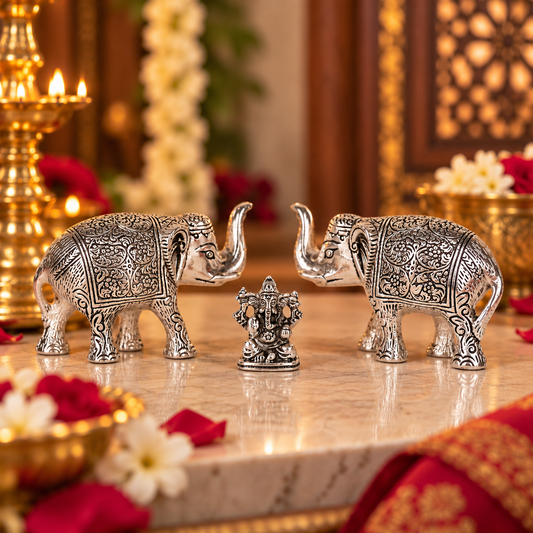 A pair of ornate silver elephant figurines facing each other with a small central Ganesha statue, displayed on a marble surface in a decorated indoor setting with red and white flowers and gold-toned decor.
