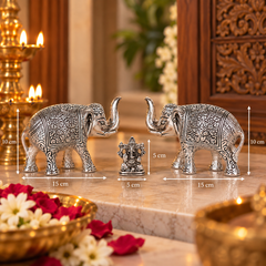 Two ornate silver elephant figurines facing each other with a small central Ganesha statue, shown on a marble surface with overlaid dimension lines indicating 15 cm length and 10 cm height. The scene is styled with lit candles, flowers, and carved wooden decor in the background.