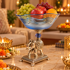 A decorative pedestal bowl with a blue translucent wavy-edged bowl and an ornate silver elephant base, filled with red apples, green grapes, and an orange, set on a marble table in a warmly lit living room.