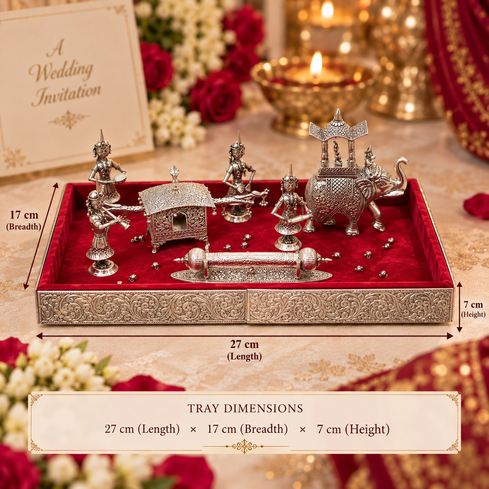 An ornate silver-toned decorative tray set displayed on a red velvet tray, with overlaid dimension labels showing 27 cm length, 17 cm breadth, and 7 cm height. The set includes multiple small metallic figurines and decorative pieces, photographed in a styled festive setting with flowers, candles, and a wedding invitation card in the background.