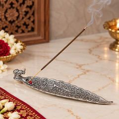 An ornate silver elephant-shaped incense holder on a marble surface, with a smoking incense stick resting across it. Decorative flowers, a carved wooden screen, and a brass lamp are visible in the background.