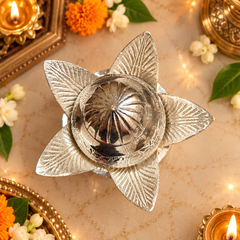 A silver-toned metal flower-shaped diya holder with a textured leaf pattern, shown on a beige surface decorated with marigolds, white flowers, and lit oil lamps.