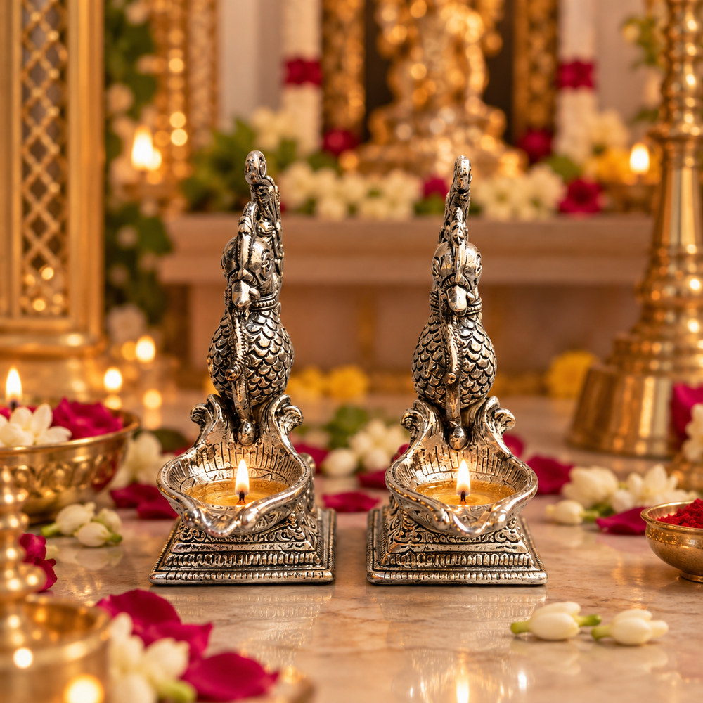 Two ornate silver-toned brass oil lamps with sculpted dragon figures on top, lit with warm flames, displayed on a marble surface with red and white flower petals and blurred golden ceremonial decor in the background.