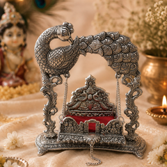 An ornate silver-toned metal Peacock Swing (Golu) idol, featuring detailed peacock carvings and a red velvet seat, displayed on a beige fabric surface with warm bokeh background lights and decorative props.