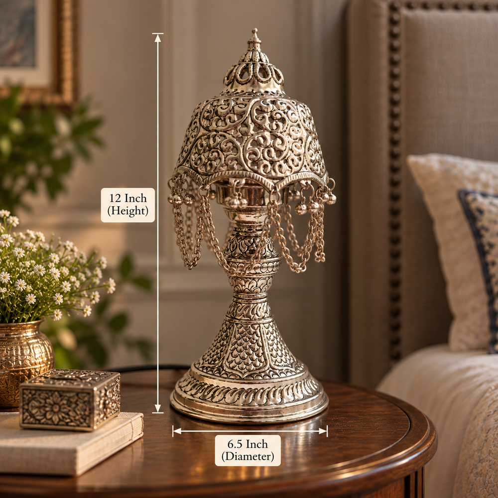 An ornate silver-toned metal table lamp with intricate filigree detailing and hanging chains, shown on a wooden table in a furnished room. White measurement lines indicate a height of 12 inches and a base diameter of 6.