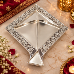 A square silver-toned decorative tray with ornate floral engraving around the border and three triangular sections, styled on a marble surface with red fabric, flowers, and candles.
