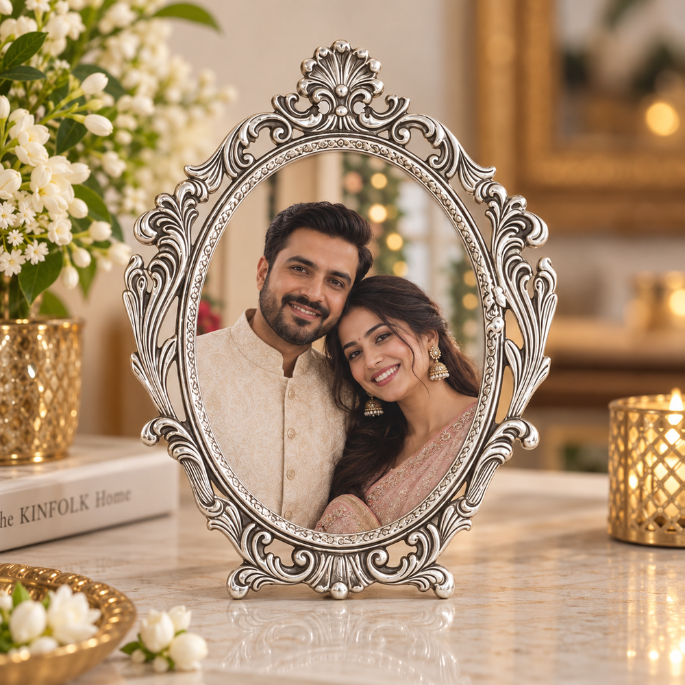 An ornate oval silver picture frame with intricate scrollwork and a small stand, holding a portrait of a smiling couple. The frame is displayed on a marble table with decorative flowers, books, and gold-toned candle holders in a warm, festive indoor setting.