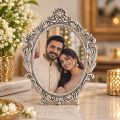 An ornate oval silver picture frame with intricate scrollwork and a small stand, holding a portrait of a smiling couple. The frame is displayed on a marble table with decorative flowers, books, and gold-toned candle holders in a warm, festive indoor setting.
