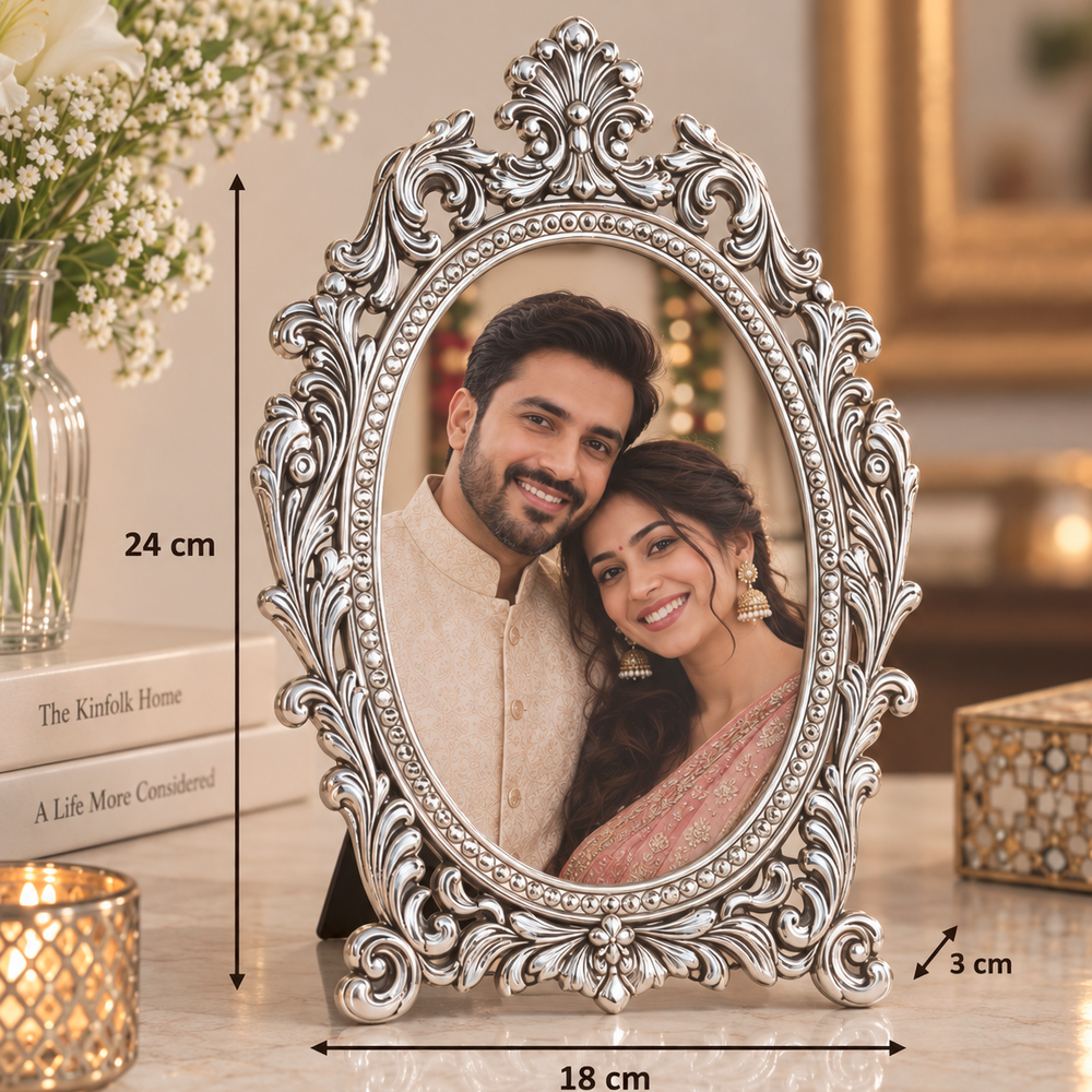 An ornate silver oval picture frame with a decorative scrollwork border, shown standing on a marble surface. The photo inside the frame depicts a smiling couple.