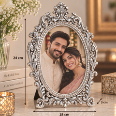 An ornate silver oval picture frame with a decorative scrollwork border, shown standing on a marble surface. The photo inside the frame depicts a smiling couple.