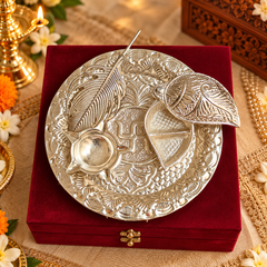 An ornate silver-toned plate with embossed floral patterns and a swastika symbol, displayed on a deep red velvet box. The plate includes a small bowl and two leaf-shaped pieces, set against a decorated festive background with flowers and candles.