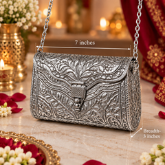 An ornate silver embossed handbag with a chain strap, shown on a marble surface with decorative flowers, candles, and red fabric in the background. White measurement lines and text indicate dimensions: 7 inches wide and 3 inches deep.