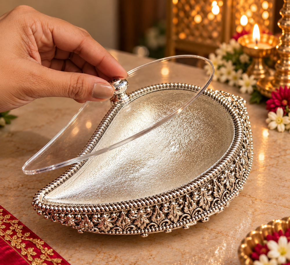 A silver teardrop-shaped decorative tray with ornate filigree detailing around the edge and a clear acrylic lid, shown on a marble surface with a hand lifting the lid. Warm candlelight and floral decorations are visible in the background.