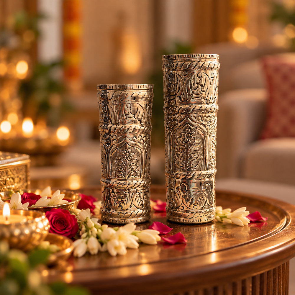Two ornate silver-toned cylindrical decorative holders with intricate embossed patterns, displayed on a wooden table in a warmly lit, festive indoor setting with flowers and candles.