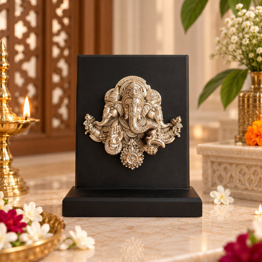 A black rectangular plaque featuring a detailed metallic Ganesha sculpture in a silver finish, displayed on a marble surface with decorative flowers and a brass lamp in a warm indoor setting.