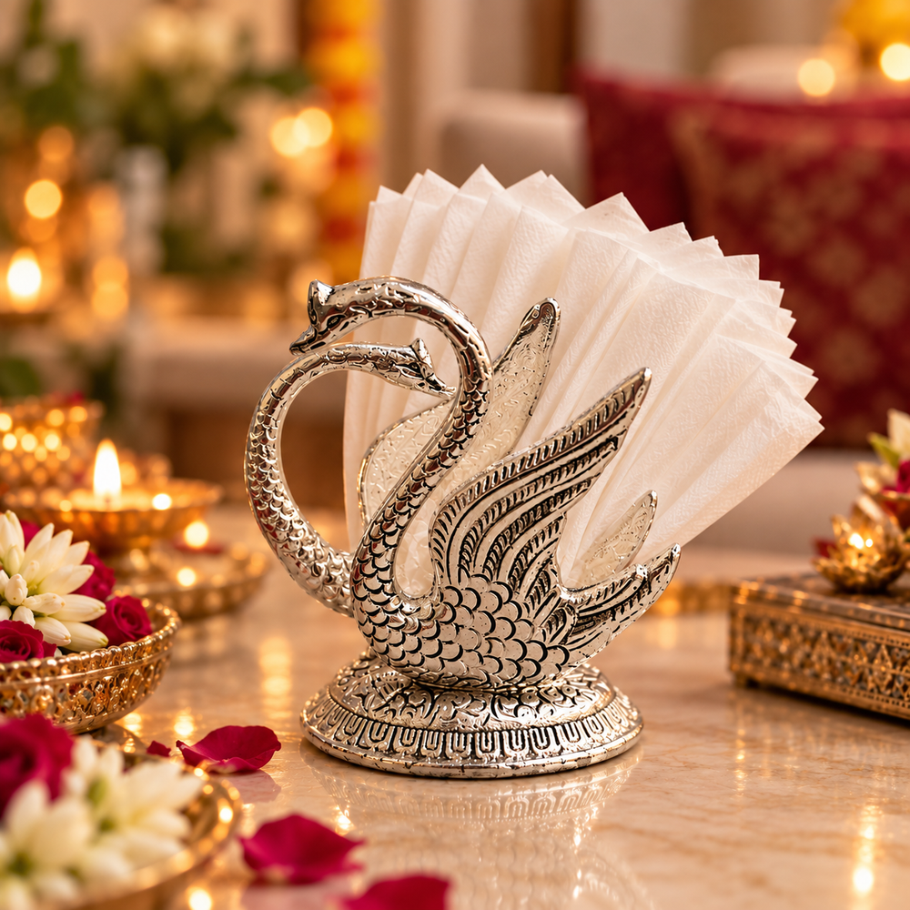An ornate silver-toned metal napkin holder shaped like two swans, holding white folded napkins, set on a decorated table with warm lights and flowers in the background.