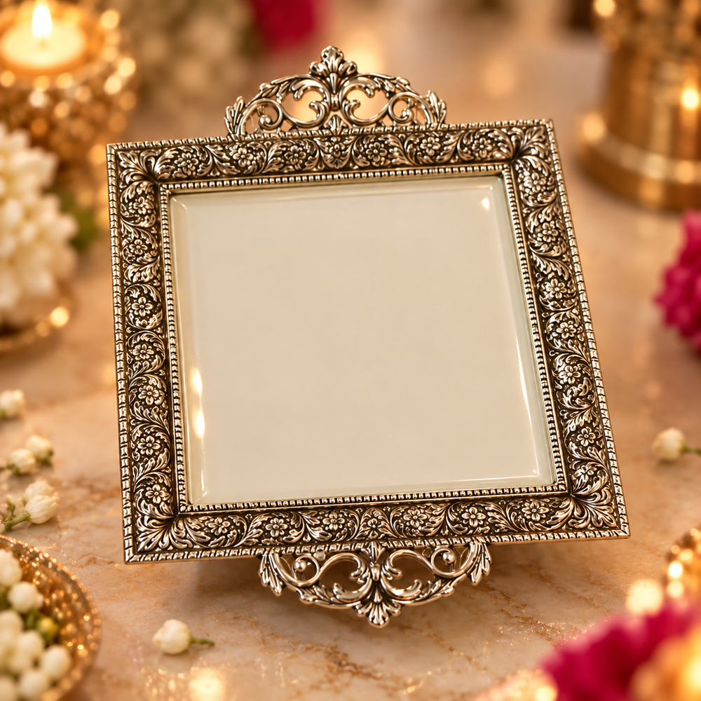 An ornate square silver-toned picture frame with intricate floral scrollwork and a decorative top and bottom crest, displayed on a marble surface with warm candlelight and flowers in the background.
