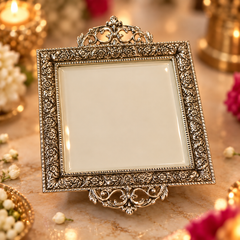 An ornate square silver-toned picture frame with intricate floral scrollwork and a decorative top and bottom crest, displayed on a marble surface with warm candlelight and flowers in the background.