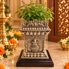 An ornate silver-toned decorative planter with carved floral details, holding green leafy stems, displayed on a dark wooden base on a marble-like surface with warm indoor decor in the background.