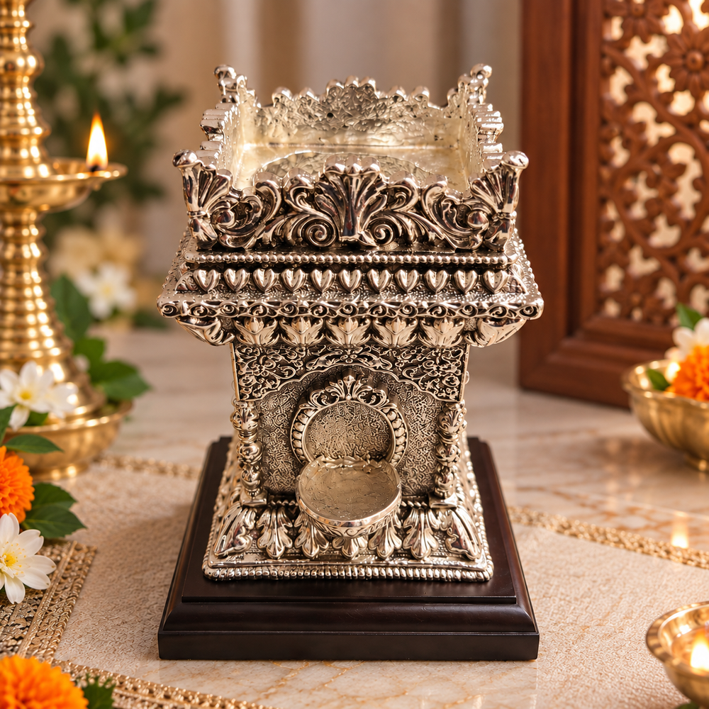 An ornate silver-toned metal oil lamp holder with intricate carvings and a small spout, displayed on a dark wooden base on a decorated table with flowers and lit candles.