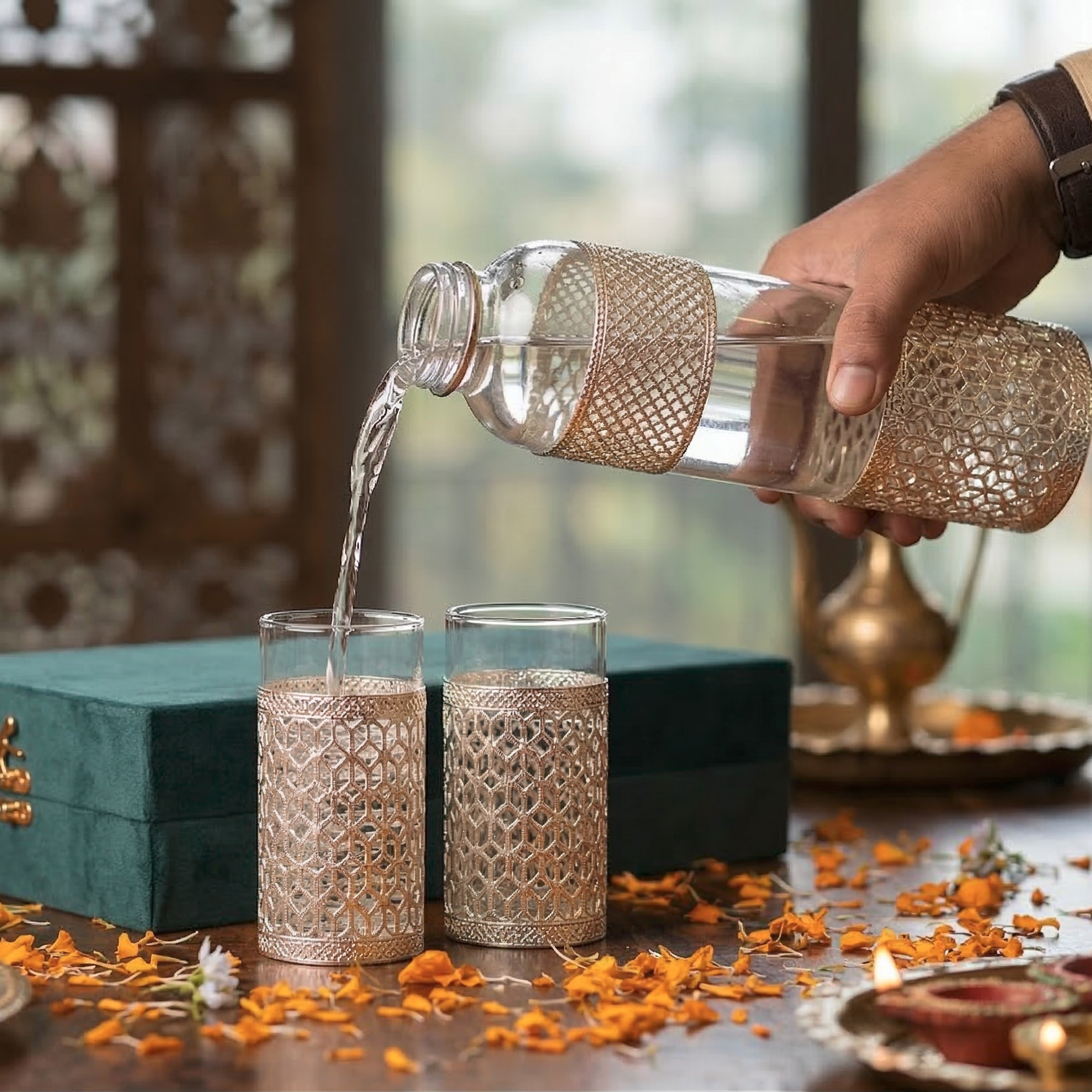 Person pouring liquid from a decorative bottle into two glasses on a table with flowers and a box.