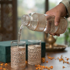 Person pouring liquid from a bottle into two ornate glasses on a wooden table with decorative elements.