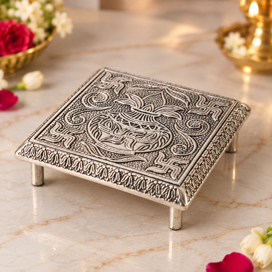 An ornate square silver-toned metal stand with intricate embossed floral and geometric patterns, shown on a polished marble surface with decorative flowers in the background.