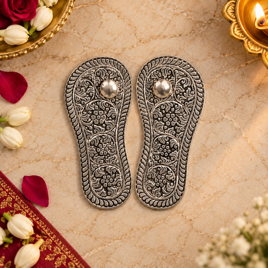A pair of ornate silver-toned flat sandals with intricate floral engravings and a small silver bead at the top, displayed on a textured beige surface with decorative flowers and a diya.