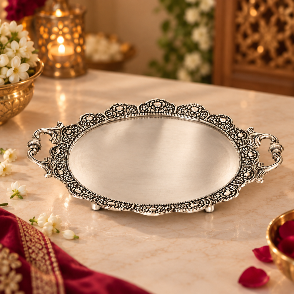 An ornate oval silver serving tray with intricate filigree borders and decorative side handles, displayed on a marble tabletop with warm decorative background elements.