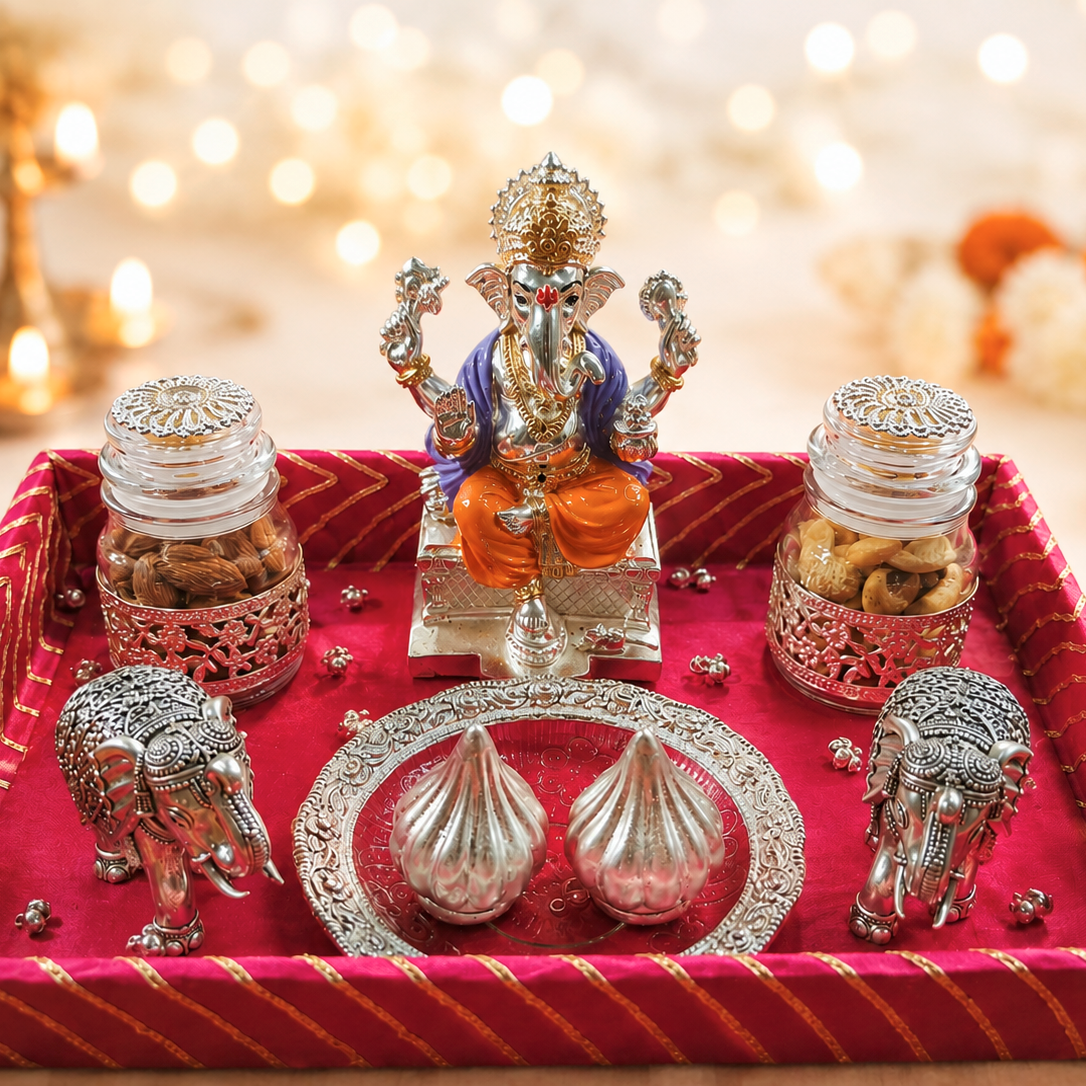 A decorative Ganesha altar tray with a silver Ganesha statue, two glass jars of sweets, silver elephant figurines, and two silver modak dishes on a red velvet tray, set against a warm bokeh background with marigolds.
