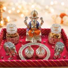 A decorative Ganesha altar tray with a silver Ganesha statue, two glass jars of sweets, silver elephant figurines, and two silver modak dishes on a red velvet tray, set against a warm bokeh background with marigolds.