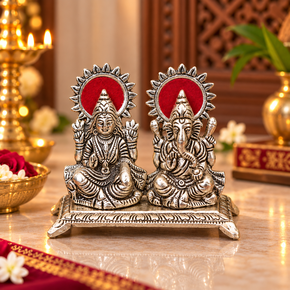 A pair of silver-toned Lord Laxmi and Ganesha idol statues with red circular backdrops, displayed on a polished marble surface with warm decorative background elements.