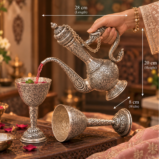 An ornate silver-toned Arabic-style teapot with intricate engraved patterns is being used to pour pink liquid into a matching engraved goblet. A second matching goblet lies on its side below.