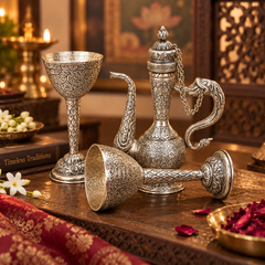 An ornate silver-toned metal tea set with intricate engraved patterns, including a tall pitcher, a goblet, and a small bowl, arranged on a wooden table in a warm, traditional setting.