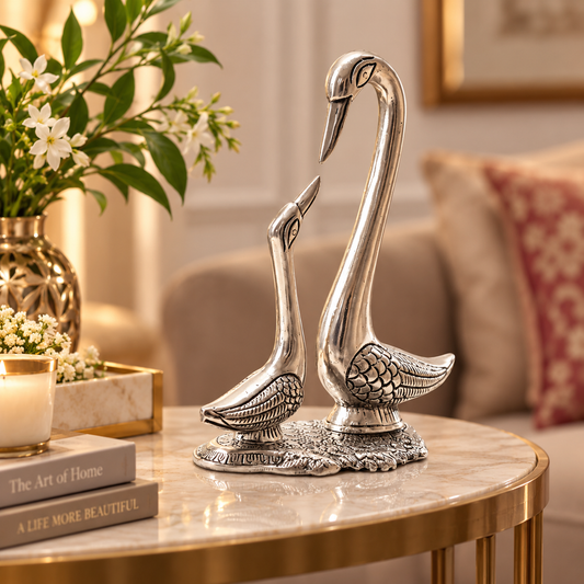 A pair of silver-toned swan figurines posed on a round marble-top table in a styled living room setting, with flowers, books, and a sofa in the background.