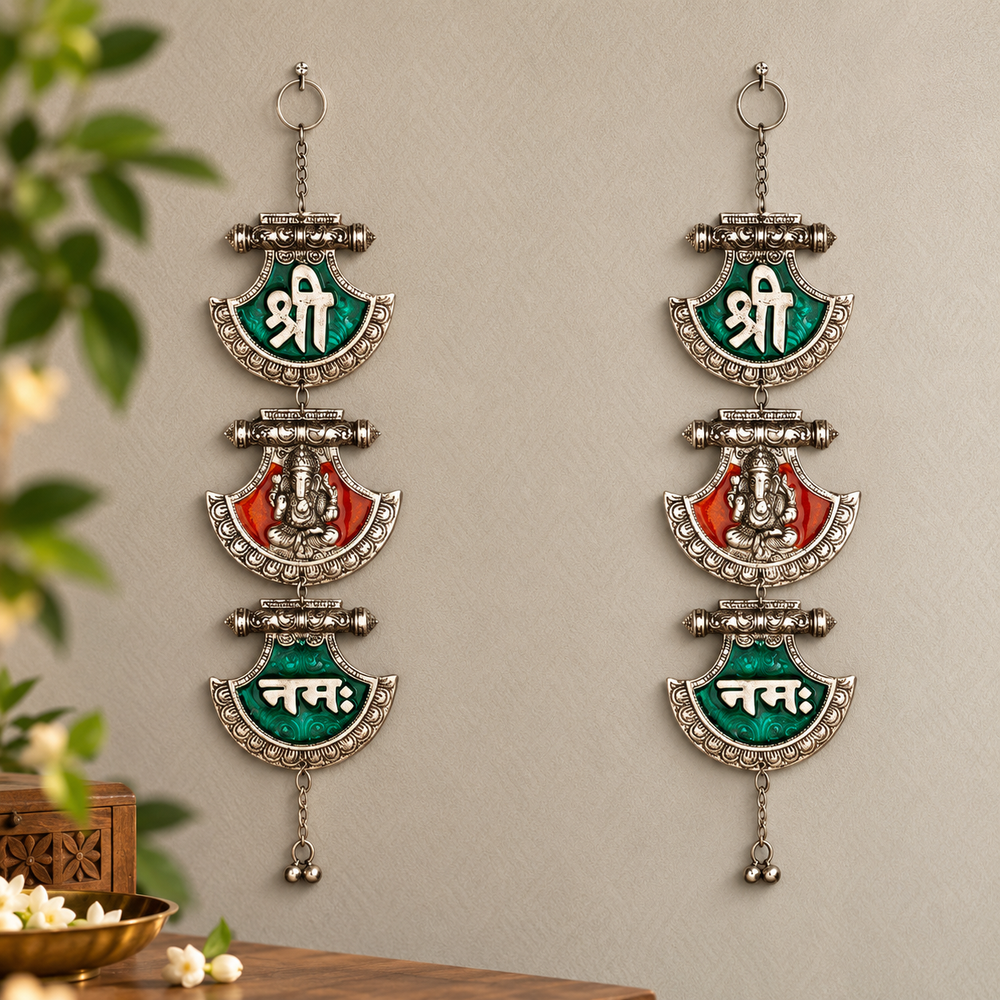 Two matching silver-toned wall hanging ornaments with green and red enamel panels and white Devanagari script, displayed against a light beige wall with decorative plants and a wooden surface at the edges.