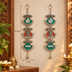 Two silver-toned hanging wall decorations with green and red enamel panels and white Devanagari text, shown on a beige wall with measurement arrows indicating 29 cm height and 7 cm width.