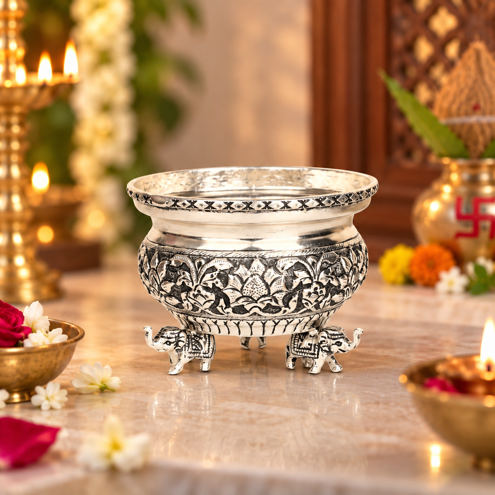 An ornate silver-toned metal pot with intricate black floral engravings and elephant feet, displayed on a marble surface with warm decorative lights and flowers in the background.